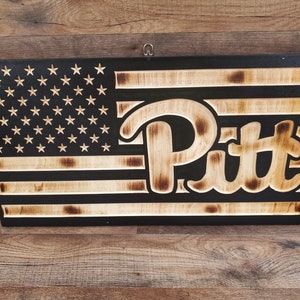 May include: A wooden American flag with the word "Pitt" burned into the wood. The flag is black with white stripes and white stars.