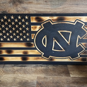 Pode incluir: Uma bandeira americana de madeira com um efeito de madeira queimada. A bandeira tem um logotipo UNC Tar Heels preto e branco no centro.