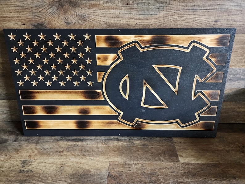 Pode incluir: Uma bandeira americana de madeira com um efeito de madeira queimada. A bandeira tem um logotipo UNC Tar Heels preto e branco no centro.