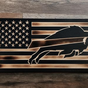 Peut inclure: Un drapeau américain en bois avec un logo de buffle noir sculpté. Le drapeau présente le design traditionnel des étoiles et des rayures, avec le logo du buffle remplaçant une partie des rayures. Le bois a une finition naturelle et brûlée.