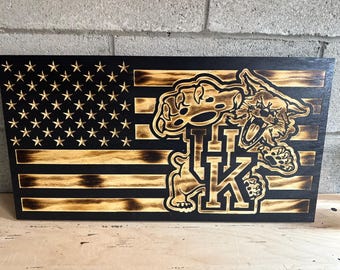 May include: Wooden American flag with a carved Kentucky Wildcats logo. The flag features the stars and stripes design, with the Wildcats logo in the center. The logo includes the letters "UK" and a wildcat.