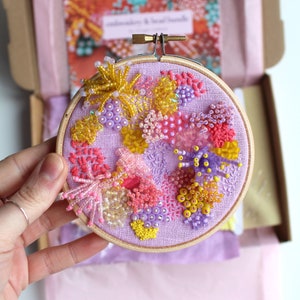 May include: A close-up of a purple embroidery hoop with a colorful beaded design. The design features pink, yellow, and purple beads arranged in a floral pattern.