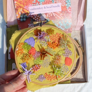 May include: A yellow embroidery hoop with a colourful floral design featuring beads and embroidery. The design includes orange, yellow, green, purple, and brown beads. The hoop is labelled "embroidery & bead bundle by imogen melissa".