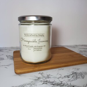 May include: A clear glass jar candle with a silver lid. The candle is labeled "Handcrafted by Stephy Honeysuckle Jasmine Soy Blend Candle with Fragrance Oil 12 oz Hand poured in California."