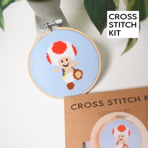 May include: Cross stitch kit featuring a pixelated image of a mushroom character from the Super Mario video game series. The kit includes a hoop, fabric, and embroidery floss. The kit is packaged in a brown cardboard box with the text "CROSS STITCH KIT" and "SUGARPLAYS" printed on it.