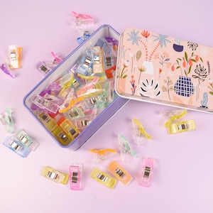 May include: An open, rectangular, light blue tin filled with colorful sewing clips, scattered on a light purple surface. The tin's lid, decorated with a floral pattern, lies open beside it. The clips are various colors.