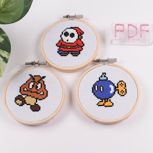 May include: Three embroidered hoop art pieces featuring characters from the Super Mario Bros. video game series. The characters are a Goomba, a Bob-omb, and a Shy Guy.