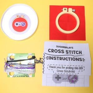 May include: A cross-stitch kit with a video game controller design. The kit includes a red fabric square, embroidery hoop, thread, and instructions. The instructions read "SUGARBPLAYS CROSS STITCH INSTRUCTIONS". The kit is on a yellow background.