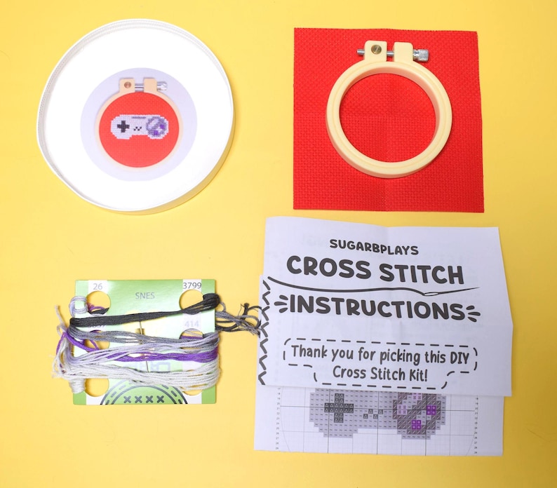 May include: A cross-stitch kit with a video game controller design. The kit includes a red fabric square, embroidery hoop, thread, and instructions. The instructions read "SUGARBPLAYS CROSS STITCH INSTRUCTIONS". The kit is on a yellow background.