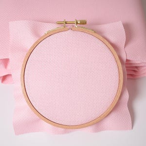 May include: A pink Aida cloth with a wooden embroidery hoop. The hoop is 8 inches in diameter.