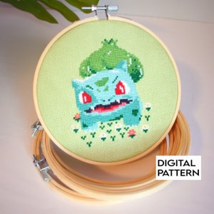 May include: A green and blue cross-stitch pattern of a Pokemon character, Bulbasaur, with a green leaf on its head. The pattern is framed in a wooden embroidery hoop.
