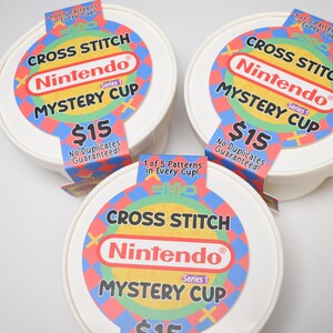 May include: Three white, round containers with colorful labels. Each label reads "CROSS STITCH Nintendo MYSTERY CUP" with the Nintendo logo. The labels also state "No Duplicates Guaranteed!" and "1 of 5 Patterns in Every Cup!"