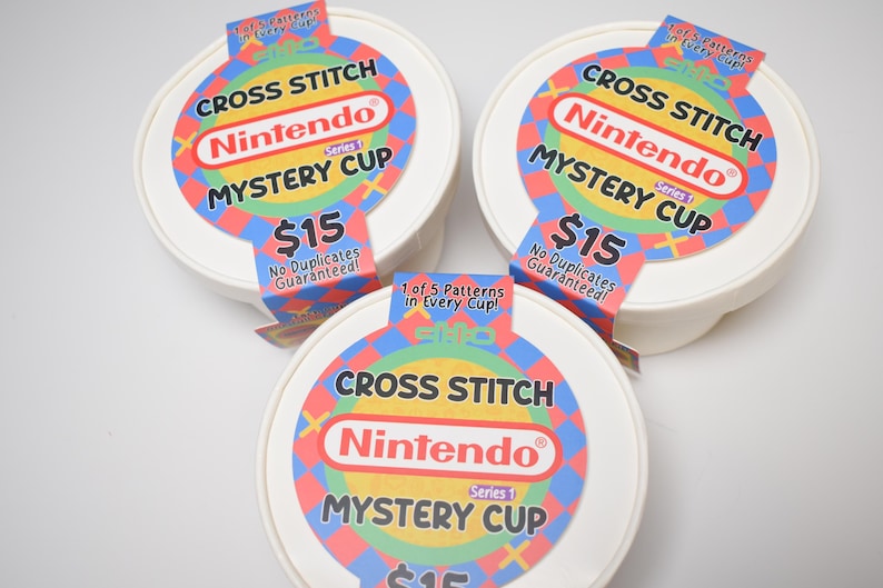 May include: Three white, round containers with colorful labels. Each label reads "CROSS STITCH Nintendo MYSTERY CUP" with the Nintendo logo. The labels also state "No Duplicates Guaranteed!" and "1 of 5 Patterns in Every Cup!"