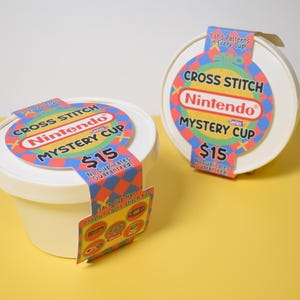 May include: Two white, round containers with lids, labeled "Cross Stitch Nintendo Mystery Cup." Each cup has a colorful label with the Nintendo logo and the text "$15." One cup has a hanging tag with images of cross-stitch patterns.