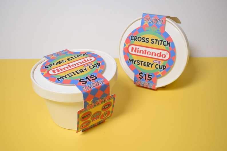 May include: Two white, round containers with lids, labeled "Cross Stitch Nintendo Mystery Cup." Each cup has a colorful label with the Nintendo logo and the text "$15." One cup has a hanging tag with images of cross-stitch patterns.