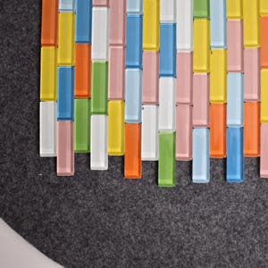 May include: A colorful mosaic tile pattern featuring rectangular glass tiles in various colors, including yellow, blue, orange, pink, green, and white. The tiles are arranged in vertical rows against a dark gray background.