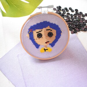 May include: A cross-stitch embroidery hoop with a purple background and a colorful image of a girl with blue hair. The girl has large eyes and a yellow shirt. The hoop is on a purple fabric surface.