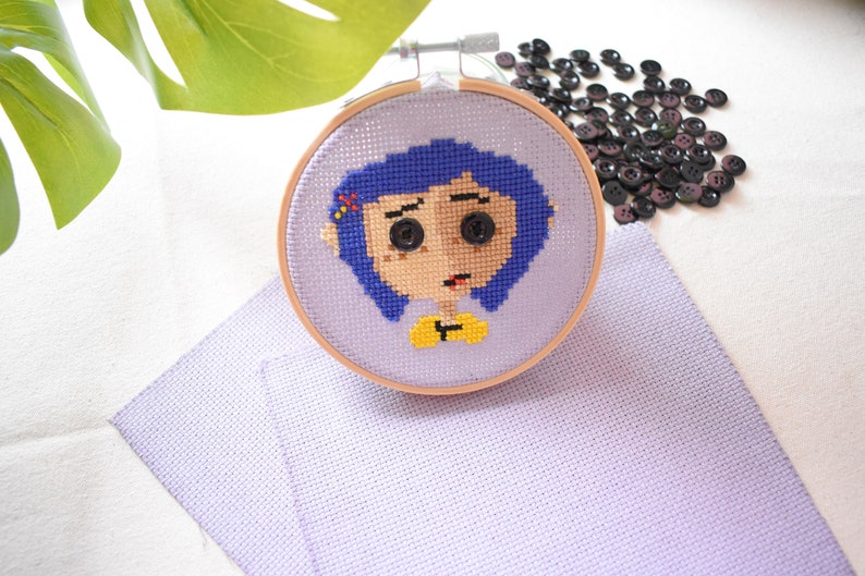 May include: A cross-stitch embroidery hoop with a purple background and a colorful image of a girl with blue hair. The girl has large eyes and a yellow shirt. The hoop is on a purple fabric surface.