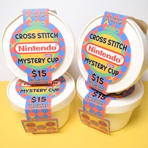 May include: Four white, round containers with lids, each labeled "Cross Stitch Nintendo Mystery Cup." The labels feature a colorful design with the Nintendo logo and the text "$15." Each cup has a tag that says "1 of 5 Patterns in Every Cup!"