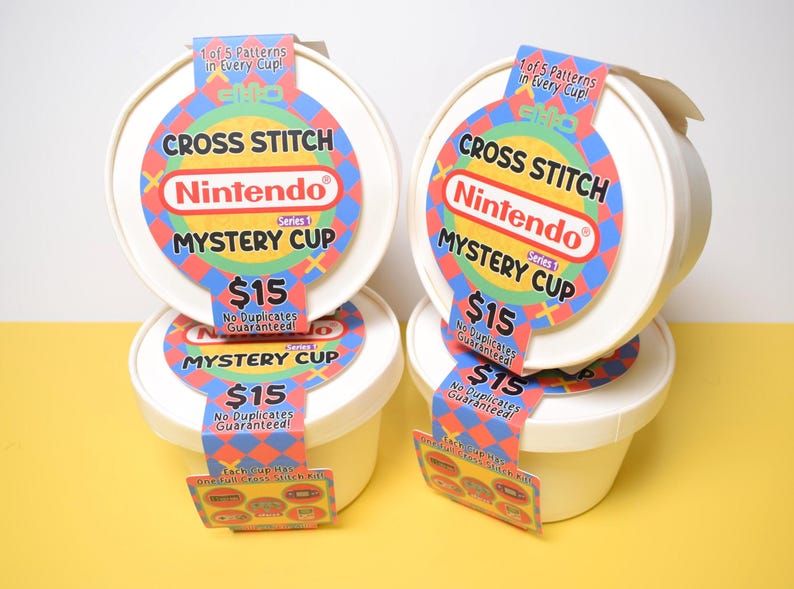 May include: Four white, round containers with lids, each labeled "Cross Stitch Nintendo Mystery Cup." The labels feature a colorful design with the Nintendo logo and the text "$15." Each cup has a tag that says "1 of 5 Patterns in Every Cup!"