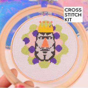 May include: Cross stitch kit featuring a colorful design of a person wearing a crown and a flower-like pattern around the head. The design is stitched on white Aida cloth and is framed in a wooden hoop.