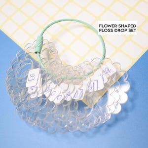 Pode incluir: Conjunto de gotas de fio transparente em forma de flor com um anel de metal verde. O conjunto inclui vários anéis de plástico transparente, cada um com uma pequena etiqueta branca com números. O texto "FLOWER SHAPED FLOSS DROP SET" é visível.