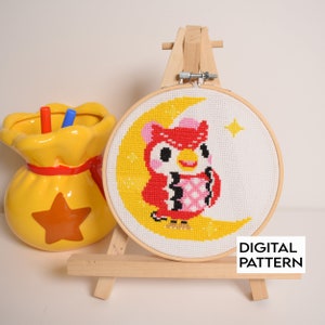 May include: A cross-stitch pattern of a red owl with a pink belly and black and white stripes on its wings, sitting on a yellow crescent moon with a yellow star in the background. The pattern is framed in a wooden hoop and displayed on a wooden easel. The text "DIGITAL PATTERN" is printed below the hoop.