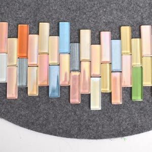 May include: An arrangement of rectangular, iridescent glass tiles in various pastel colors, including pink, orange, blue, and green. The tiles are arranged on a gray felt surface, creating a colorful mosaic pattern.