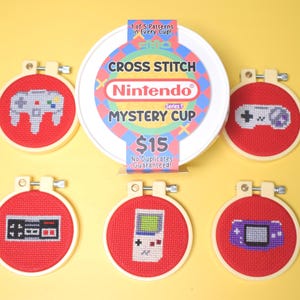May include: A collection of cross-stitch embroidery hoops featuring video game controller designs on red fabric. The hoops are displayed with a white container labeled "Cross Stitch Nintendo Mystery Cup". The text "1 of 5 Patterns in Every Cup" is also visible.