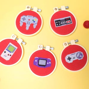 May include: Five red cross-stitch hoops with video game controller designs. The designs include a classic controller, a handheld console, and a retro game controller. The hoops are set against a yellow background with red felt balls.