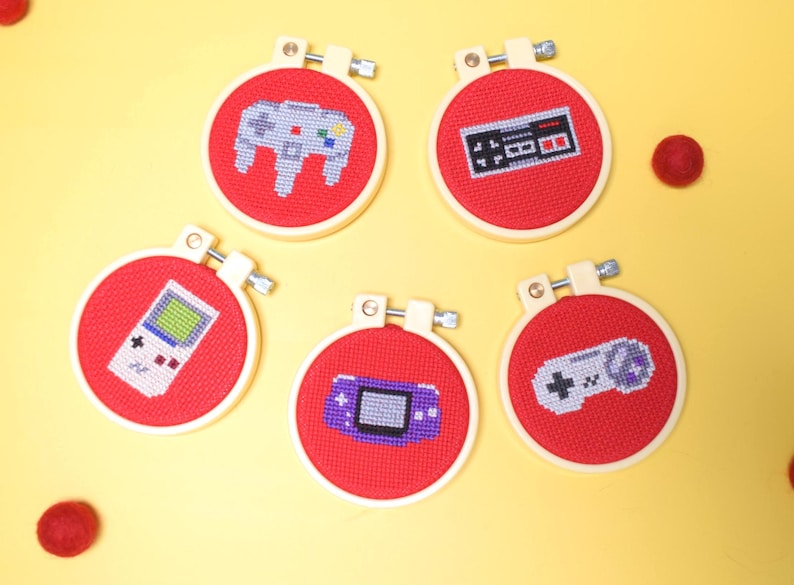 May include: Five red cross-stitch hoops with video game controller designs. The designs include a classic controller, a handheld console, and a retro game controller. The hoops are set against a yellow background with red felt balls.