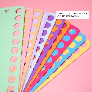 May include: A fan of colorful thread organizer cards in various shades, including green, pink, orange, purple, blue, and yellow. Each card features a series of hexagonal cutouts and a heart-shaped hole. The text "THREADS ORGANIZER CARD (10 PACK)" is visible.