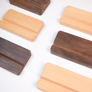 May include: A collection of wooden phone stands in various shades of brown and natural wood. Each stand has a rectangular shape with a slot for holding a phone. The stands are arranged on a white surface.
