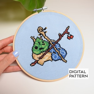 May include: Cross-stitch embroidery hoop with a blue background featuring a green and brown creature with a blue and white bag and a brown stick with a red berry. The text "DIGITAL PATTERN" is at the bottom of the hoop.