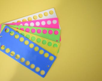 Könnte beinhalten: Eine Sammlung farbenfroher Maschenhalter aus Kunststoff in Blau, Gelb, Grün, Pink und Weiß, jeweils mit gleichmäßig verteilten kreisförmigen Löchern. Die Maschenhalter sind auf einem gelben Hintergrund angeordnet.