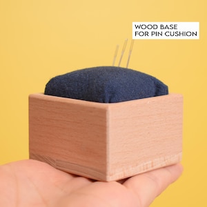 Könnte beinhalten: Ein quadratisches Nadelkissen mit einem dunkelblauen Stoffoberteil in einem hellen Holzsockel. Drei dünne Nähnadeln aus Metall stecken im Stoff. Der Text "WOOD BASE FOR PIN CUSHION" ist in Schwarz auf einem weißen rechteckigen Etikett aufgedruckt.