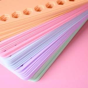 K&ouml;nnte beinhalten: Ein Stapel bunter, dreieckiger Kunststofftabletts in Pastellt&ouml;nen von Pfirsich, Rosa, Blau, Lila und Gr&uuml;n. Jedes Tablett hat sechseckige Aussparungen. Die Tabletts sind auf einem rosa Hintergrund gestapelt.