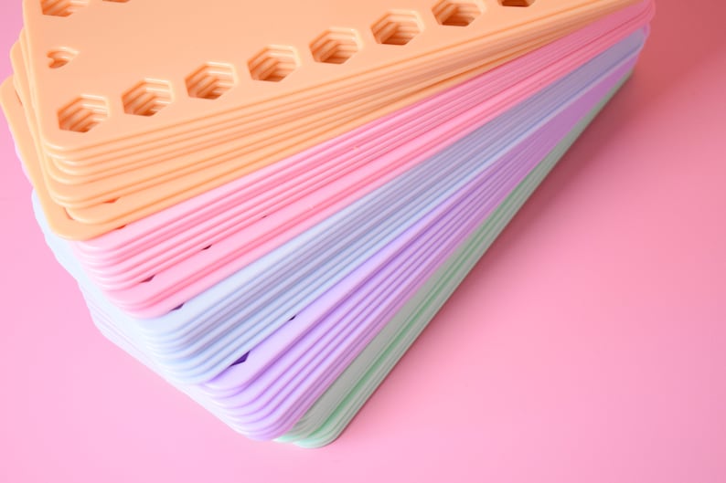 K&ouml;nnte beinhalten: Ein Stapel bunter, dreieckiger Kunststofftabletts in Pastellt&ouml;nen von Pfirsich, Rosa, Blau, Lila und Gr&uuml;n. Jedes Tablett hat sechseckige Aussparungen. Die Tabletts sind auf einem rosa Hintergrund gestapelt.