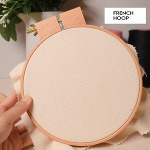 Puede incluir: Un aro de bordado de madera con una tela beige estirada sobre él. El aro se sostiene en una mano y descansa sobre una superficie de tela beige. El texto "FRENCH HOOP" es visible en la esquina superior derecha de la imagen.