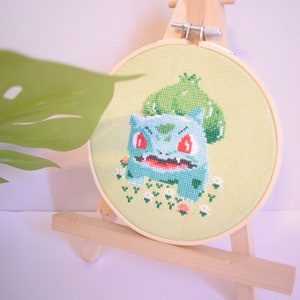 K&ouml;nnte beinhalten: Ein gestickter Kreuzstich-Hoop mit einem Cartoon-Bild von Bisasam, einem Pok&eacute;mon-Charakter, mit roten Augen und einem gr&uuml;nen, bl&auml;ttrigen Kopf. Der Hoop steht auf einem Holzst&auml;nder.