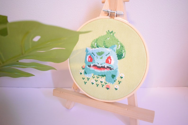 K&ouml;nnte beinhalten: Ein gestickter Kreuzstich-Hoop mit einem Cartoon-Bild von Bisasam, einem Pok&eacute;mon-Charakter, mit roten Augen und einem gr&uuml;nen, bl&auml;ttrigen Kopf. Der Hoop steht auf einem Holzst&auml;nder.