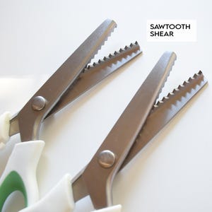 Pode incluir: Close-up de duas tesouras serrilhadas com cabos brancos e lâminas prateadas. As lâminas têm uma borda serrilhada. O texto "SAWTOOTH SHEAR" está em uma caixa branca no canto superior direito.