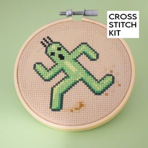 May include: Cross stitch kit featuring a green pixelated character running and leaving a trail of brown pixels behind it. The design is on a beige fabric and is framed in a wooden hoop.