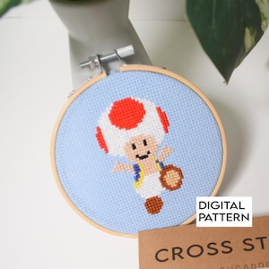 May include: A cross-stitch project in a round wooden embroidery hoop featuring a pixelated Toad character with a red and white mushroom cap, blue vest, and brown shoes on a light blue fabric background, with the text DIGITAL PATTERN visible.