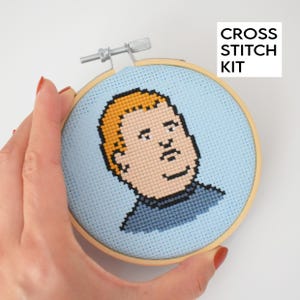 May include: A cross stitch kit featuring a cartoon portrait of a person with blonde hair and a blue shirt. The image is stitched on light blue fabric and framed in a wooden hoop. The text "CROSS STITCH KIT" is in the upper right corner.