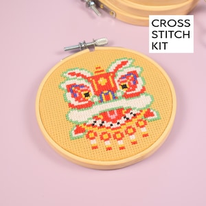 May include: A cross stitch kit featuring a colorful lion dance design. The embroidery is set in a wooden hoop, with the text "CROSS STITCH KIT" in a white box. The lion dance design includes red, green, yellow, and white colors.