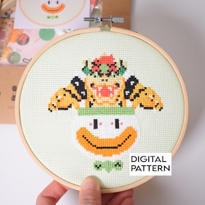 以下が含まれることがあります： 黄色とオレンジの Bowser のキャラクターが緑と白の Boo のコスチュームを着ているクロスステッチパターン。パターンは木製の刺繍枠に収められています。枠の下には「DIGITAL PATTERN」というテキストが印刷されています。