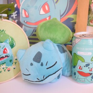 K&ouml;nnte beinhalten: Ein gr&uuml;nes und blaues Pl&uuml;sch-Bulbasaur-Spielzeug, ein gr&uuml;nes und blaues Kreuzstichbild von Bulbasaur und eine blaue und gr&uuml;ne Dose mit einem Bulbasaur-Motiv und dem Text "Ocean Bomb" darauf.