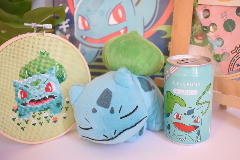 K&ouml;nnte beinhalten: Ein gr&uuml;nes und blaues Pl&uuml;sch-Bulbasaur-Spielzeug, ein gr&uuml;nes und blaues Kreuzstichbild von Bulbasaur und eine blaue und gr&uuml;ne Dose mit einem Bulbasaur-Motiv und dem Text "Ocean Bomb" darauf.