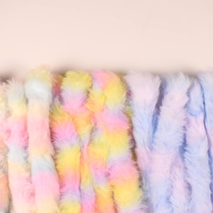 May include: Close-up of several strands of fluffy, faux fur in pastel colors. The strands are vertically oriented and display a gradient of pink, yellow, and blue hues. The soft texture and vibrant colors suggest a craft or textile product.